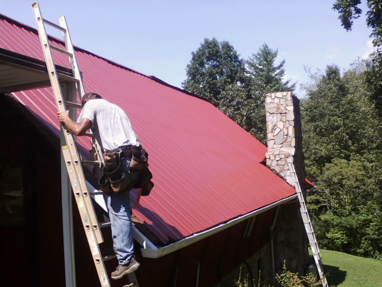 Roofer on a ladder beside a red metal roof and stone chimney — Soffit & Fascia Repair by Roofing Brothers in Neilton